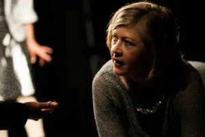 Actor in grey sparkle top and necklace kneeling, looking upward toward another performer out of frame, dramatic stage lighting