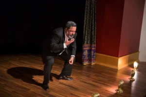 Actor in black suit crouching on wooden stage, one hand raised to chest, near ornate red pillar with gold trim