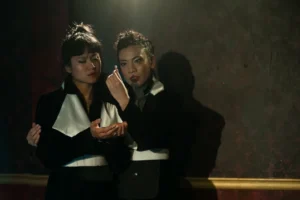Two actors in black-and-white costumes in dramatic stage lighting, one holding a small object near the other's face