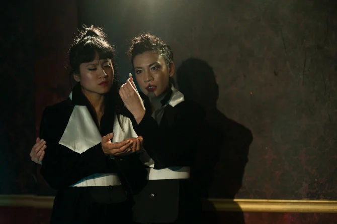 Two actors in black-and-white costumes in dramatic stage lighting, one holding a small object near the other's face