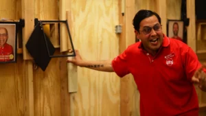 Actor in red uniform gesturing animatedly beside a wall-mounted display in a wooden set