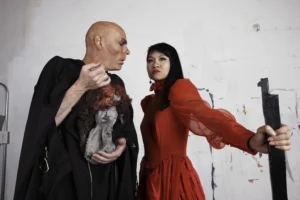 Two actors in theatrical scene: bald figure in black robe holds tattered doll; woman in red gown grips dark post