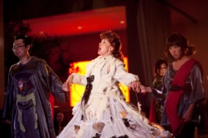 Actors in costume on stage; center performer in white fur gown sings dramatically, flanked by robed cast members